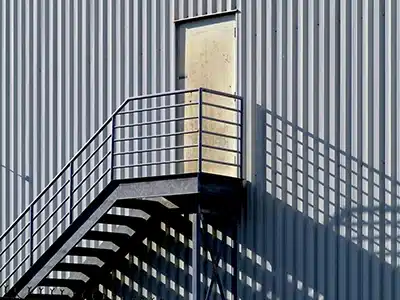Panel metálico con diseño de líneas verticales, ideal para fachadas industriales. Se observa una escalera metálica y una puerta de acceso en altura, reflejando la funcionalidad y resistencia del material.